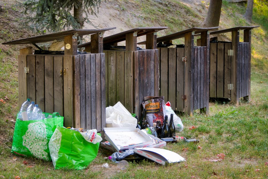 Four wooden outdoor rubbish bins with sloped roofs are positioned side by side on a grassy area, each fitted with metal hinges and locks. The bins appear weathered, with visible wood grain and a dark 